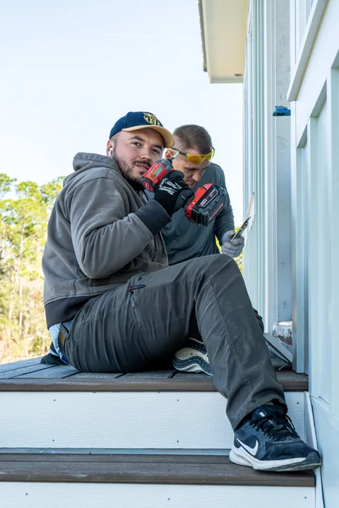 Two workers on exterior steps beside white siding, one holding a cordless drill.