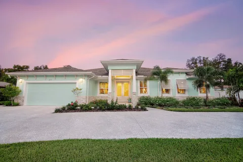 Pale turquoise single-story home with white trim, an illuminated entrance and palm trees beneath a pink evening sky.