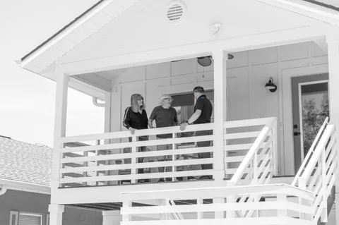 Three adults talking on the porch of an elevated home in a mostly black-and-white photograph.