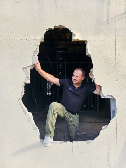 A man in a Polo Builders shirt stepping through an opening in a wall at a construction site.