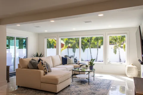 Living room with a cream sectional, blue pillows, a round glass table and windows facing palm trees.